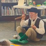 Hal Pratt dressed as a leprechaun sitting cross-legged on the floor of the library, holding a wooden mallet and speaking to a group of children. Behind him are bookshelves and a table with props including a jug and small figurines.