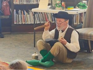 Hal Pratt dressed as a leprechaun sitting cross-legged on the floor of the library, holding a wooden mallet and speaking to a group of children. Behind him are bookshelves and a table with props including a jug and small figurines.
