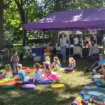 A lively outdoor storytime session under a purple tent. A group of children sit on colorful mats and cushions, attentively listening to a storyteller with open books in hand. Other people are browsing library information booths in the background.