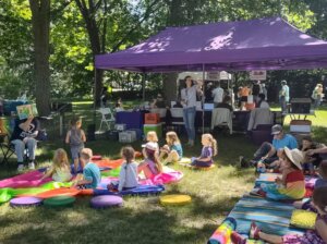 A lively outdoor storytime session under a purple tent. A group of children sit on colorful mats and cushions, attentively listening to a storyteller with open books in hand. Other people are browsing library information booths in the background.