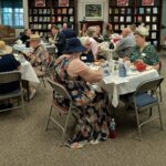 A cozy tea party setup inside a library. Guests wearing festive attire sit at tables adorned with floral tablecloths and tea sets, enjoying tea and conversation. Bookshelves filled with books line the walls.