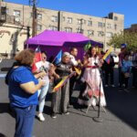 A festive outdoor event featuring a group of people holding various country flags and microphones, participating in a cultural celebration. A woman in a traditional dress leads the group while standing near a purple tent. A crowd and urban buildings form the backdrop.