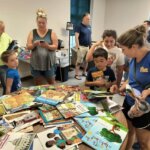 A table covered with a large selection of colorful children's books, with adults and children gathered around looking at and selecting books in a bright library space.