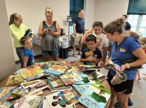 A table covered with a large selection of colorful children's books, with adults and children gathered around looking at and selecting books in a bright library space.