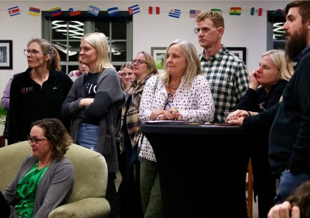 A group of adults standing attentively in a library setting, with international flags displayed above them. Some individuals are seated in the foreground, while others are standing near high-top tables, engaged in listening.