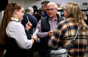 A young woman dressed as a reporter engaging in a conversation with two adults at the Murder Mystery event. They appear to be discussing or sharing information in a lively social setting.