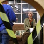 A theatrical scene with a woman sitting on a couch wearing gloves, framed by caution tape. She is a docent at the Murder Mystery event, with another person standing nearby, partially visible.