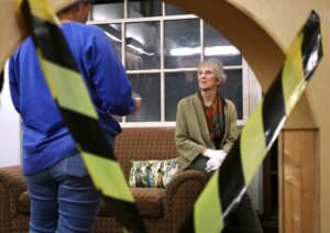 A theatrical scene with a woman sitting on a couch wearing gloves, framed by caution tape. She is a docent at the Murder Mystery event, with another person standing nearby, partially visible.