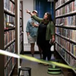 A woman pointing towards a shelf in a library aisle while holding a booklet, with a man standing behind her also holding a booklet. The scene is framed by bookshelves on both sides, and caution tape partially crosses the foreground. From the library's Murder Mystery event.