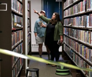 A woman pointing towards a shelf in a library aisle while holding a booklet, with a man standing behind her also holding a booklet. The scene is framed by bookshelves on both sides, and caution tape partially crosses the foreground. From the library's Murder Mystery event.