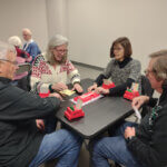 Four adults are seated around a table playing bridge. They are engaged with cards, score pads, and card holders labeled "WBC." The setting appears to be a casual gathering in a well-lit room with additional tables and participants visible in the background. The atmosphere is focused and friendly.