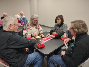 Four adults are seated around a table playing bridge. They are engaged with cards, score pads, and card holders labeled "WBC." The setting appears to be a casual gathering in a well-lit room with additional tables and participants visible in the background. The atmosphere is focused and friendly.