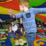 Children play under a colorful parachute during a group activity on a vibrant alphabet rug, laughing and enjoying the moment.