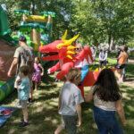 Families and children enjoy a sunny day in a park, interacting with inflatable play structures and a person in a red dragon costume.