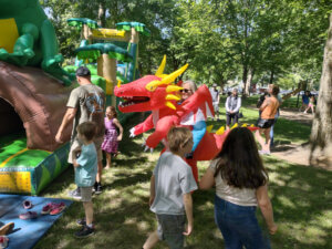 Families and children enjoy a sunny day in a park, interacting with inflatable play structures and a person in a red dragon costume.