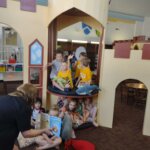 A group of children and Ms. Angie gather inside a castle-themed reading area in the library, with kids holding books and listening to a story.