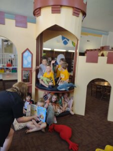 A group of children and Ms. Angie gather inside a castle-themed reading area in the library, with kids holding books and listening to a story.