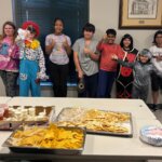 A group of children and teens pose for a photo in a room with large windows and framed artwork on the walls. One person is dressed as a clown in a colorful costume, while others wear casual clothes or costumes. In the foreground, a table is set with trays of nachos, cheese, salsa, and other snacks. The atmosphere suggests a festive gathering or event.