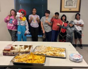 A group of children and teens pose for a photo in a room with large windows and framed artwork on the walls. One person is dressed as a clown in a colorful costume, while others wear casual clothes or costumes. In the foreground, a table is set with trays of nachos, cheese, salsa, and other snacks. The atmosphere suggests a festive gathering or event.