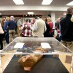 A library exhibit featuring artifacts like a cat mummy and statues is on display, with a crowd of people mingling in the background.