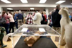 A library exhibit featuring artifacts like a cat mummy and statues is on display, with a crowd of people mingling in the background.
