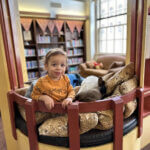 A toddler wearing an orange shirt stands in a cozy reading nook, surrounded by cushions in a library setting with shelves of books and a couch in the background.