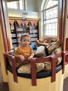 A toddler wearing an orange shirt stands in a cozy reading nook, surrounded by cushions in a library setting with shelves of books and a couch in the background.