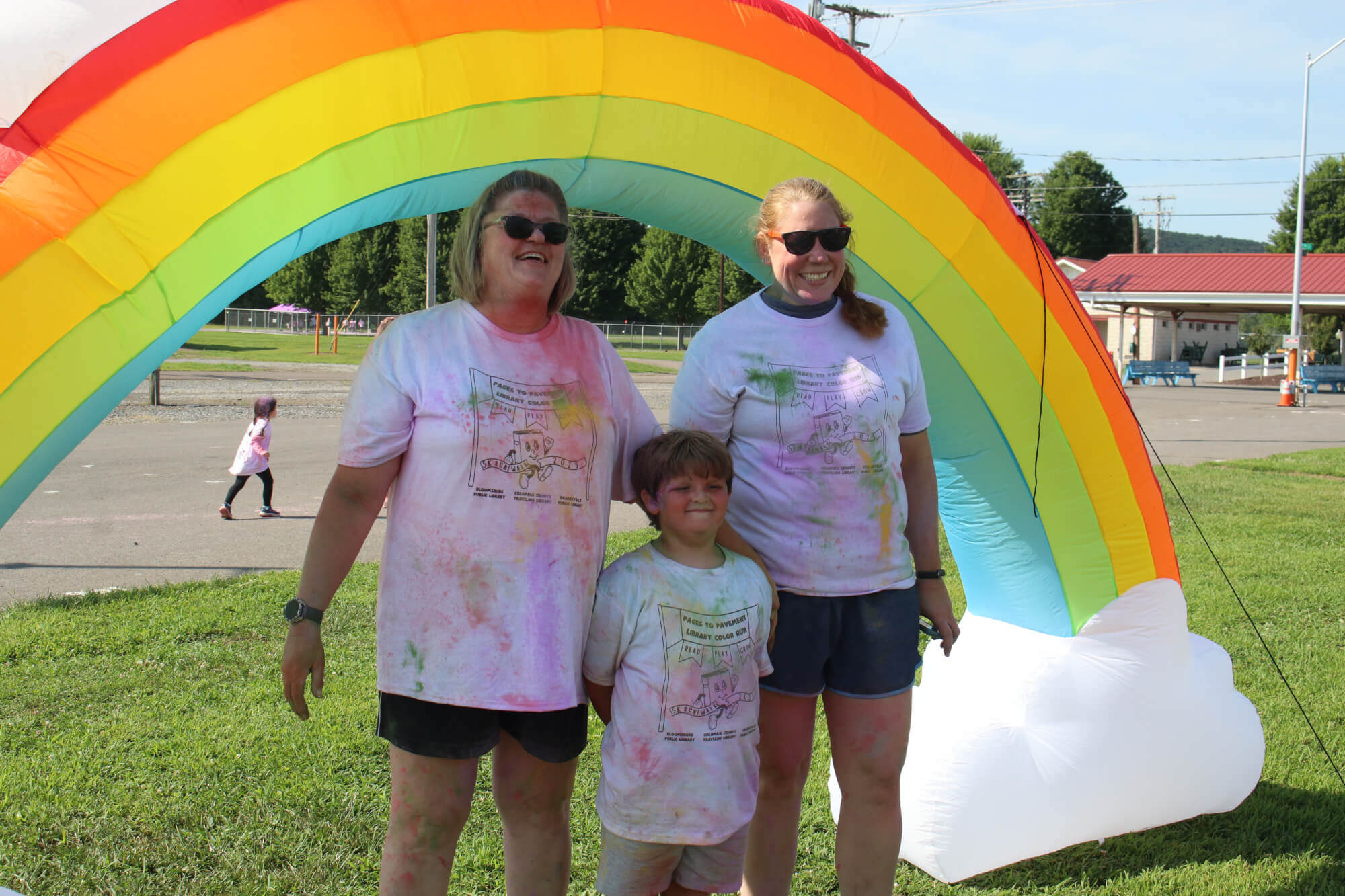 Two adults and one child with color powder splashed on their white t-shirts under an inflatable rainbow.